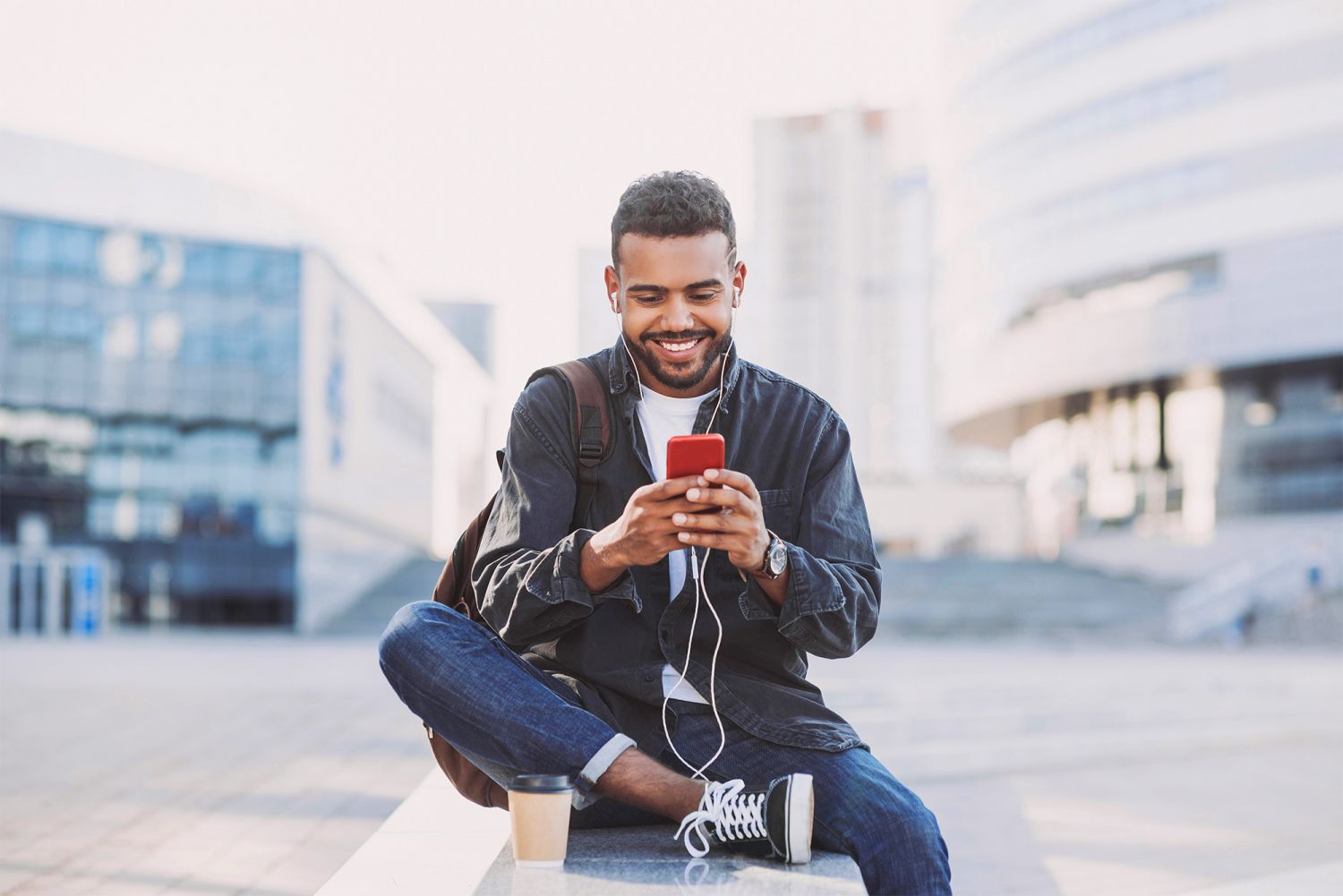 Junger Mann sitzt draußen und schaut lächelnd auf sein Handy